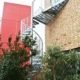 external spiral staircase with a large triangular platform, plants below and a red building behind.