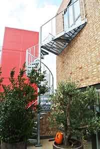 external spiral staircase with a large triangular platform, plants below and a red building behind.
