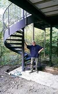man leaning against a dark steel spiral staircase. Woodland setting