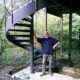 man leaning against a dark steel spiral staircase. Woodland setting